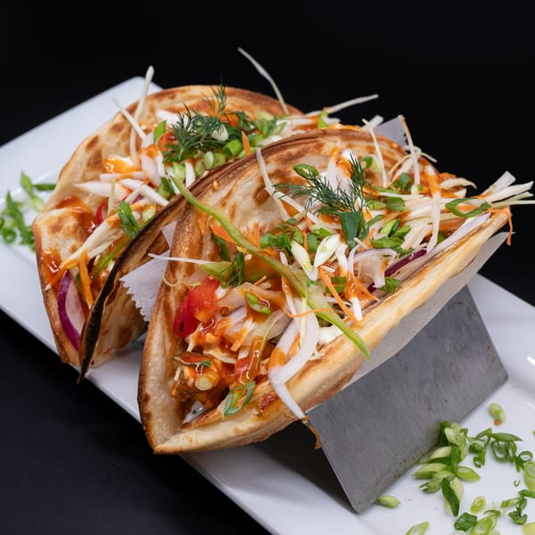 Two Indian tacos, filled with shredded vegetables and sauce, on a white plate with a black background.