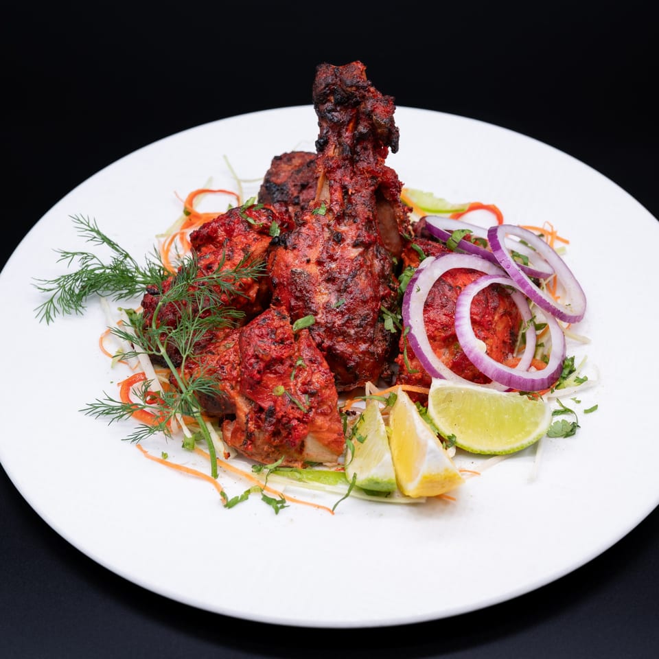 Ambrosia Zaika's Indian Tandoori Chicken, garnished with red onions, lime, and fresh dill on a white plate.
