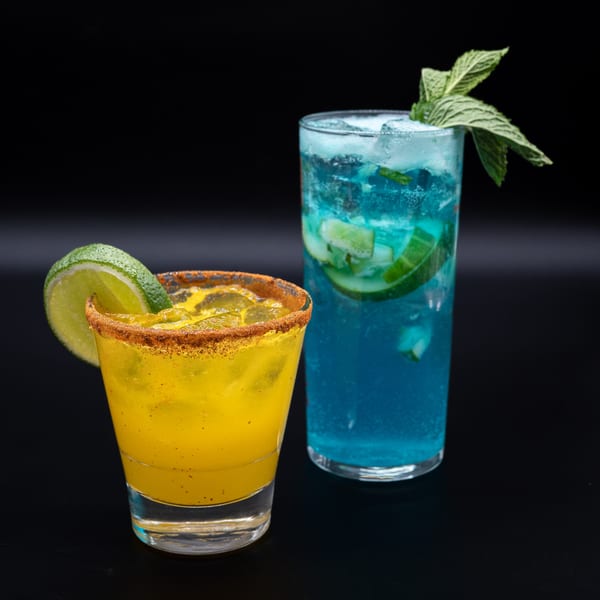 Two vibrant cocktails: a yellow drink with a lime and spiced rim, and a blue drink with mint and cucumber.